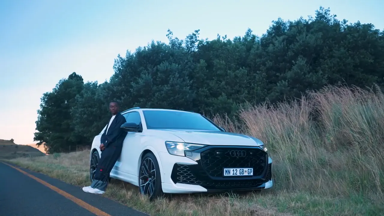 Presenter leaning on a white Audi RSQ8 parked in tall grass beside the road